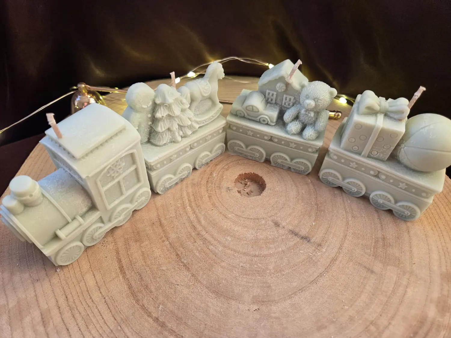 All four Christmas train candles lined up on a wood slice, fairy lights behind