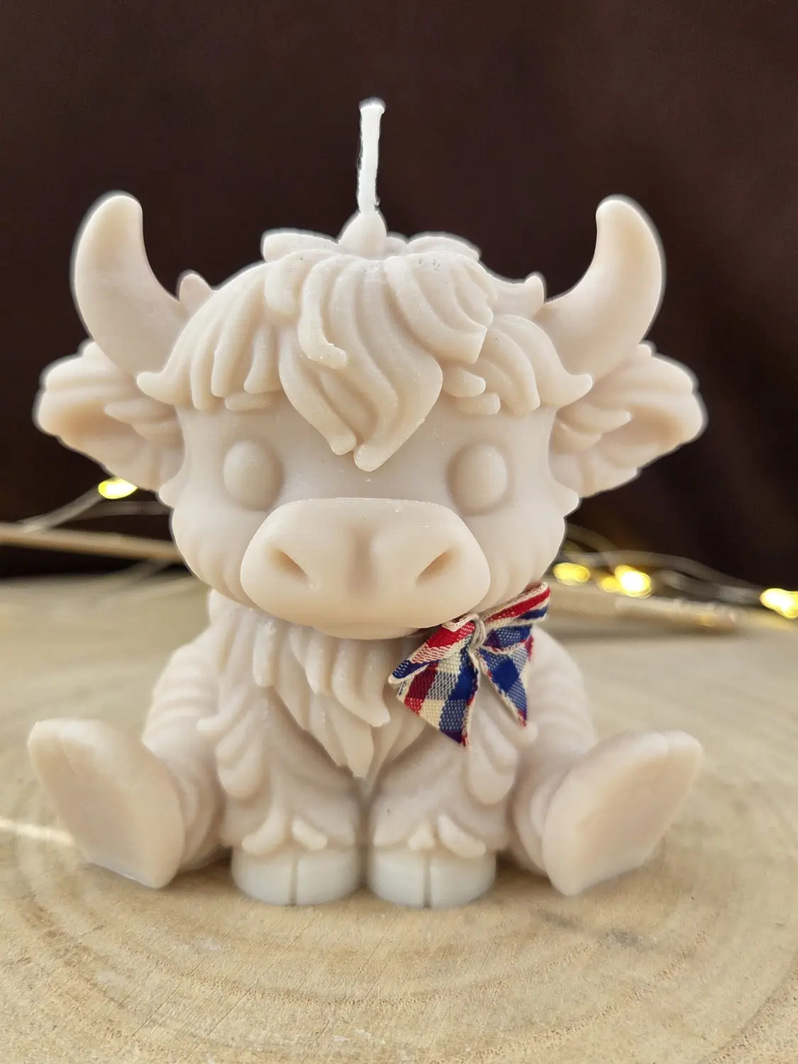Pale pink Highland cow candle with a red, white and blue checked bow, sitting on a wood slice
