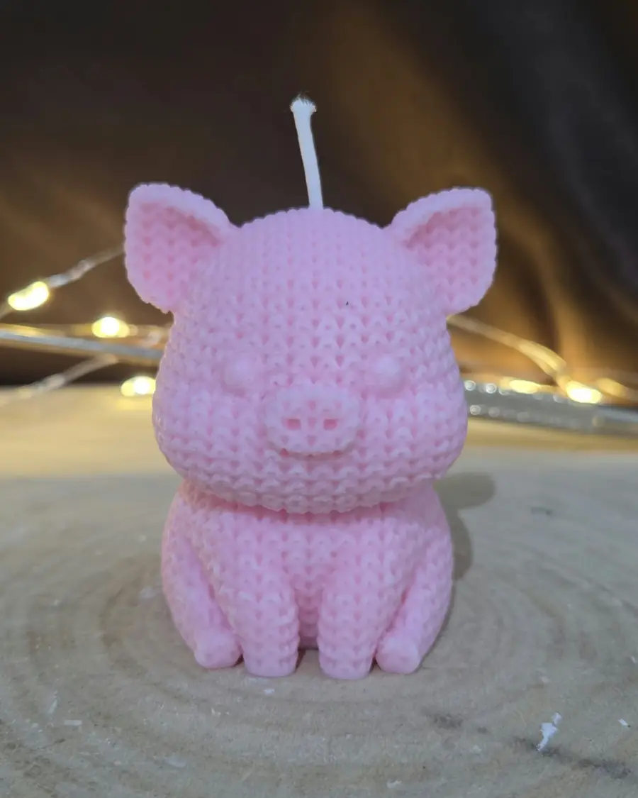 Pink pig candle with a knitted texture, sitting up on a wood slice with fairy lights behind