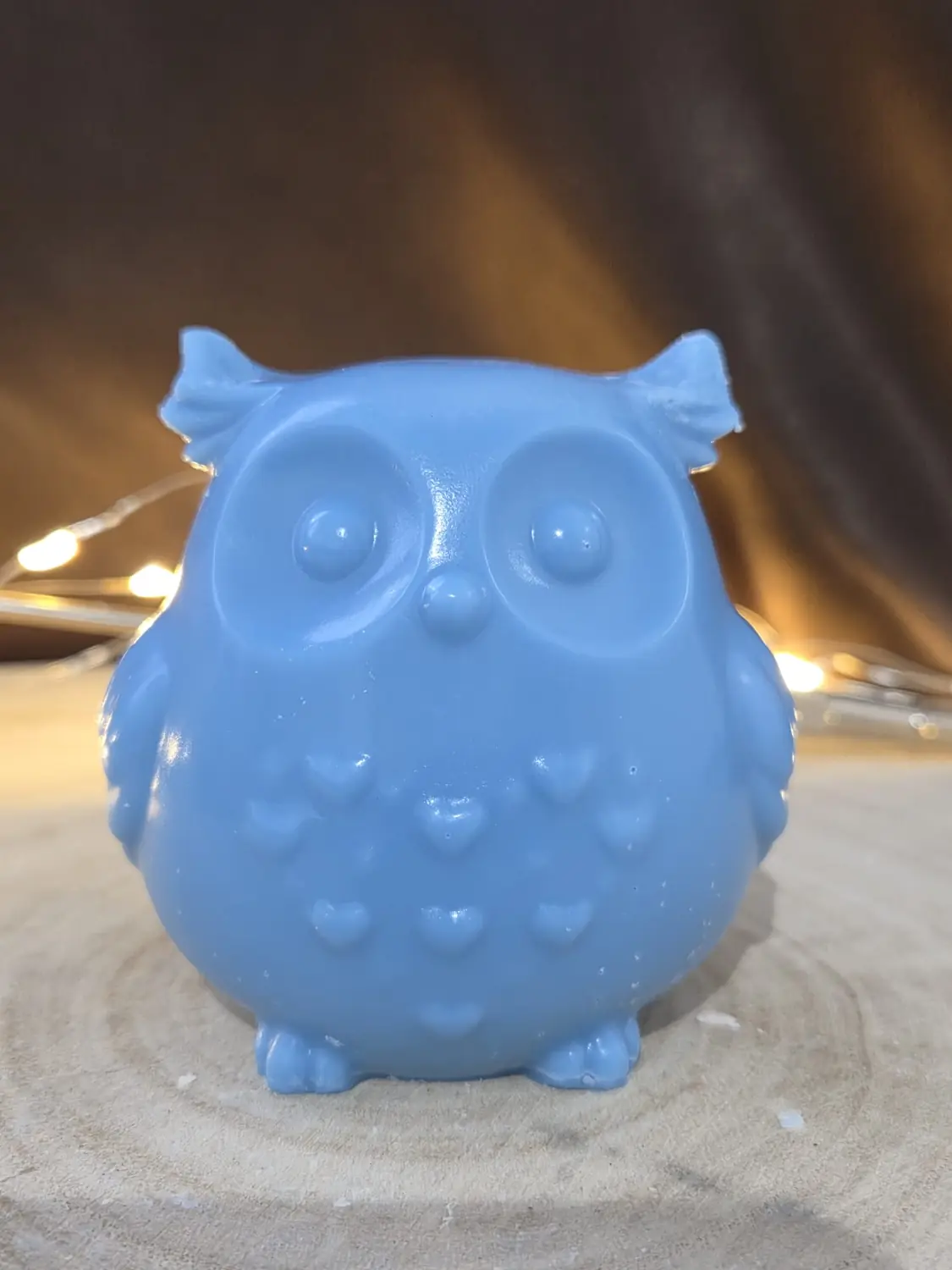 Blue owl candle with big round eyes and hearts on its chest, on a wood slice