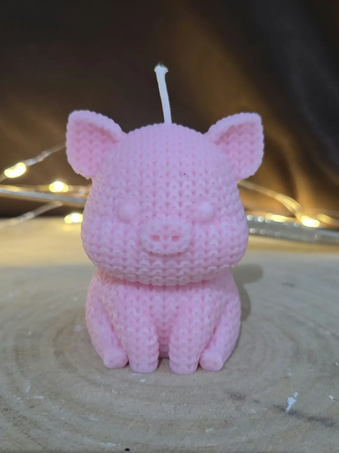 Pink pig candle with a knitted texture, sitting up on a wood slice with fairy lights behind